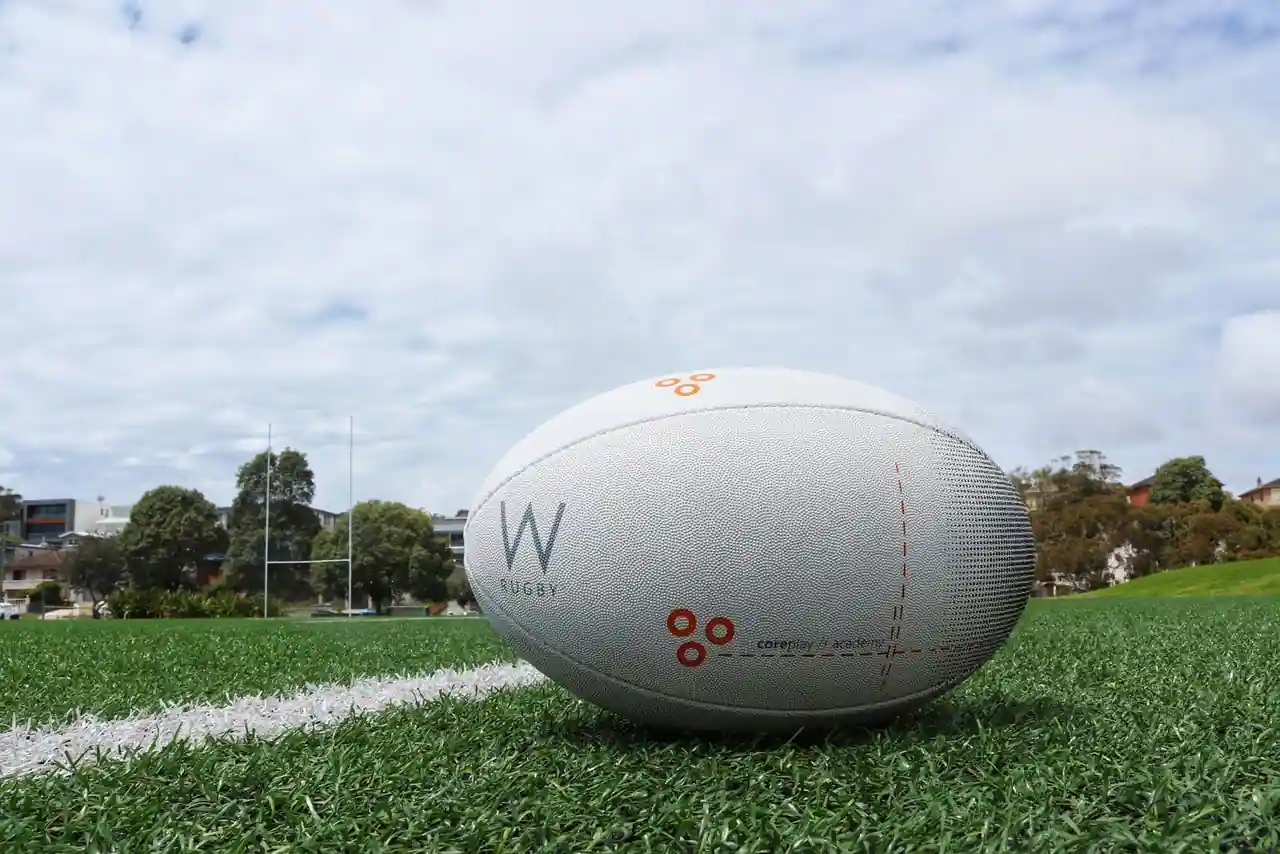 Un ballon de rugby blanc posé sur la pelouse d'un terrain, poteaux visibles en arrière-plan.