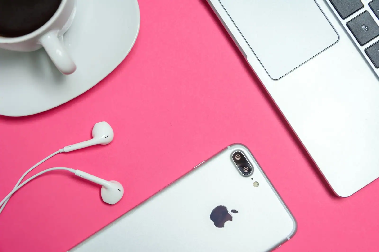 Smartphone de la marque Apple, écouteurs filaires et coin d'un ordinateur portable sur fond rose.