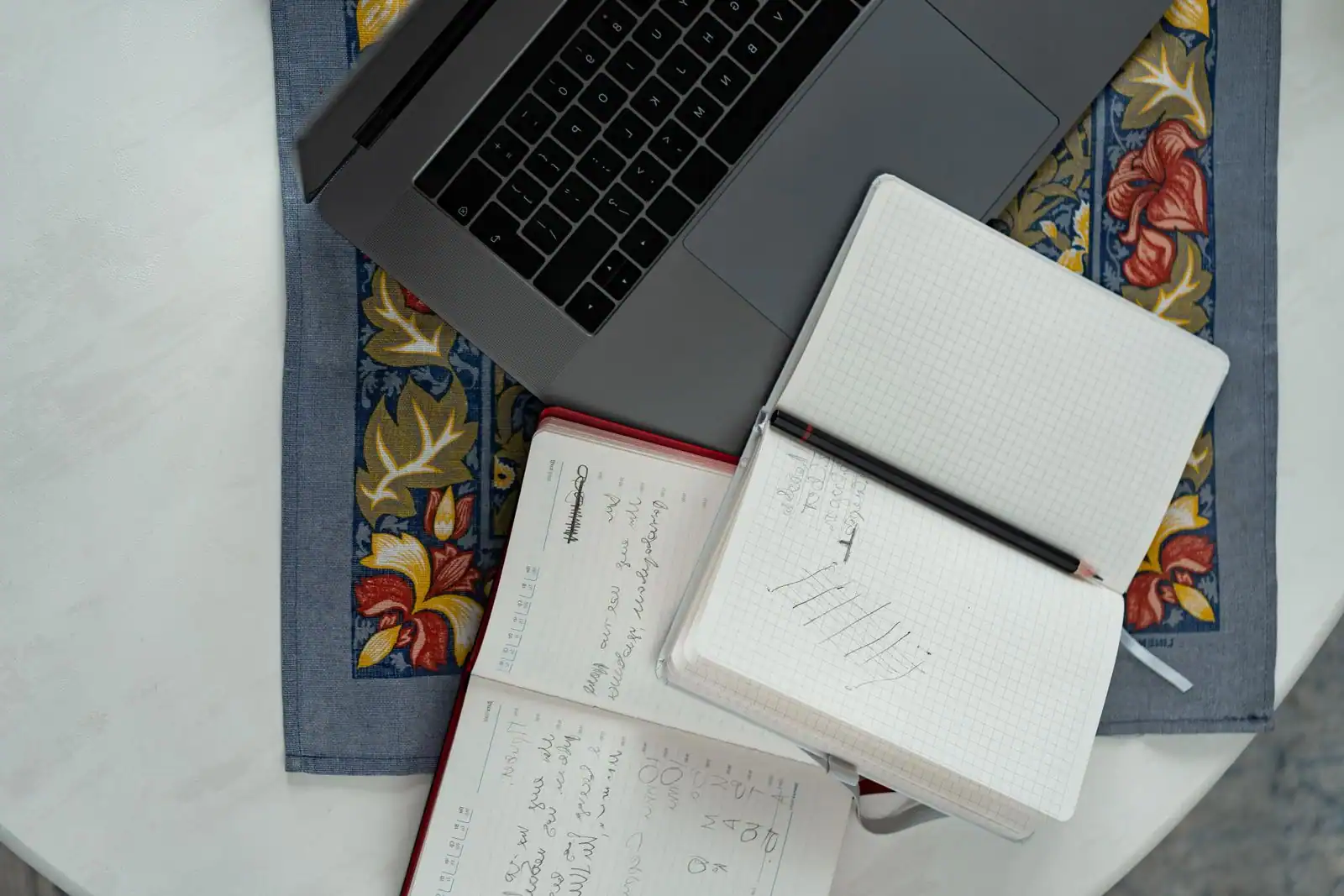 Carnet à petits carreaux et cahier de notes manuscrites posés à côté d'un ordinateur portable sur une table.