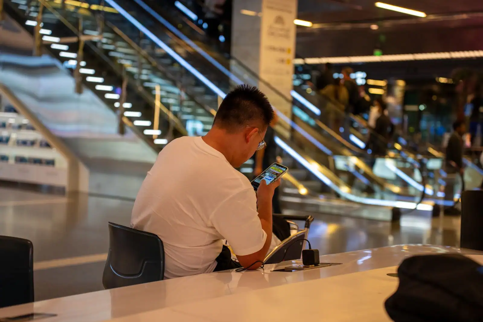Voyageur assis dans le hall d'un aéroport, consultant son smartphone près des escalators.