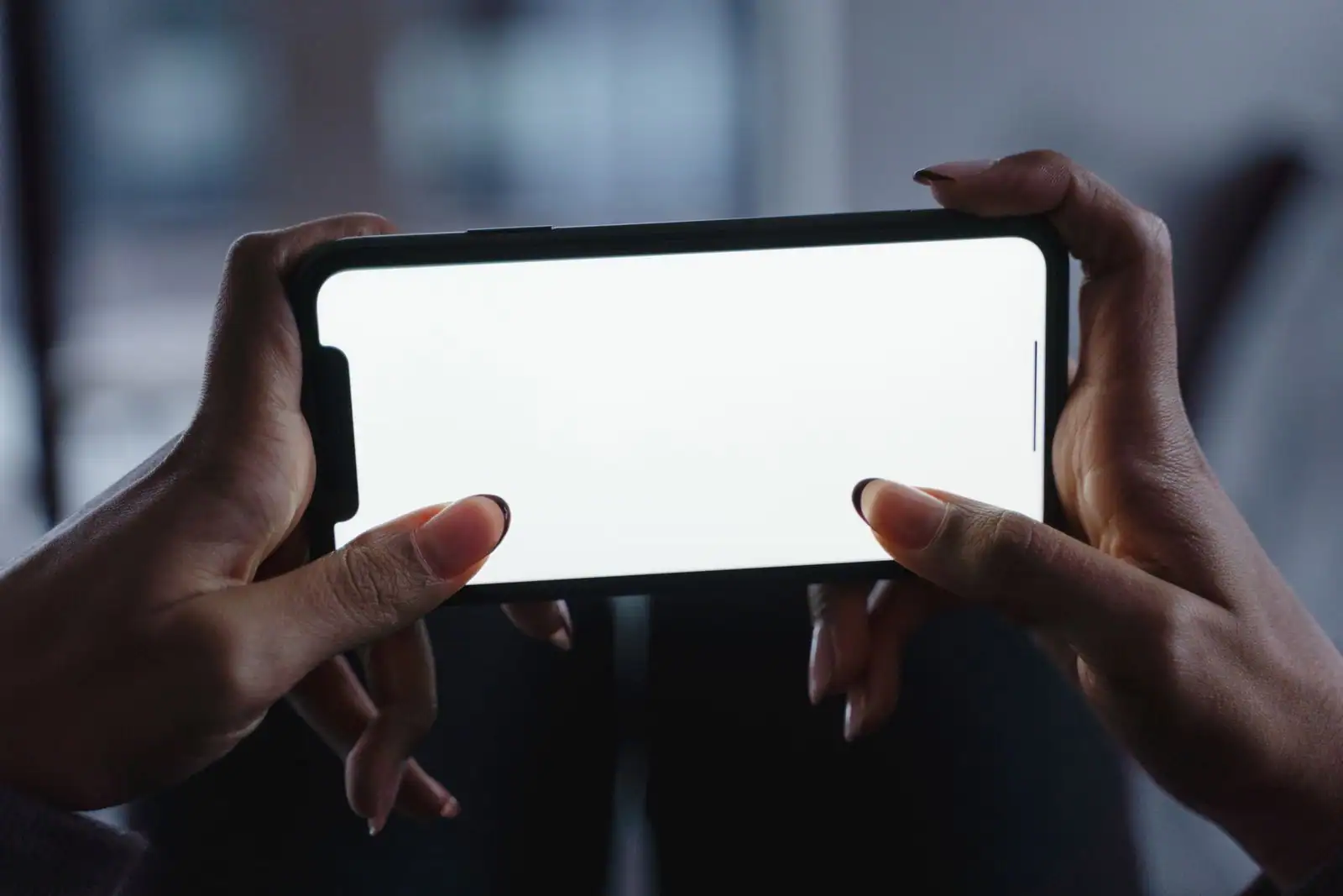 Mains tenant un smartphone à l'écran blanc allumé, dans une pièce sombre.