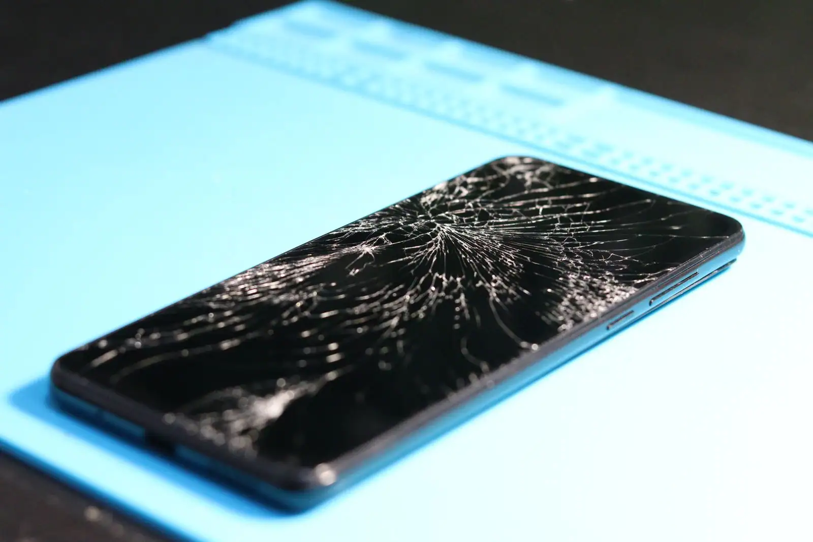 Smartphone à l'écran fortement fissuré et brisé, posé sur une surface bleue de réparation.