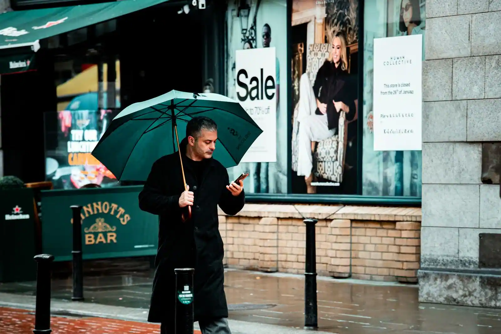 Homme sous un parapluie consultant son smartphone dans une rue détrempée par la pluie.