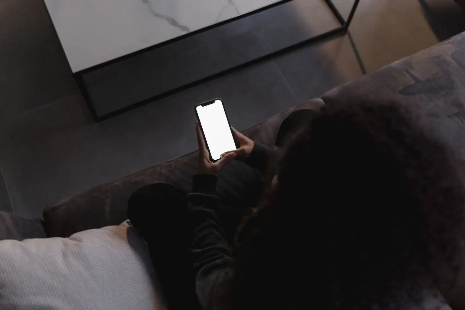 Personne assise sur un canapé tenant un smartphone à l'écran vierge, sans marque visible sur l'appareil.
