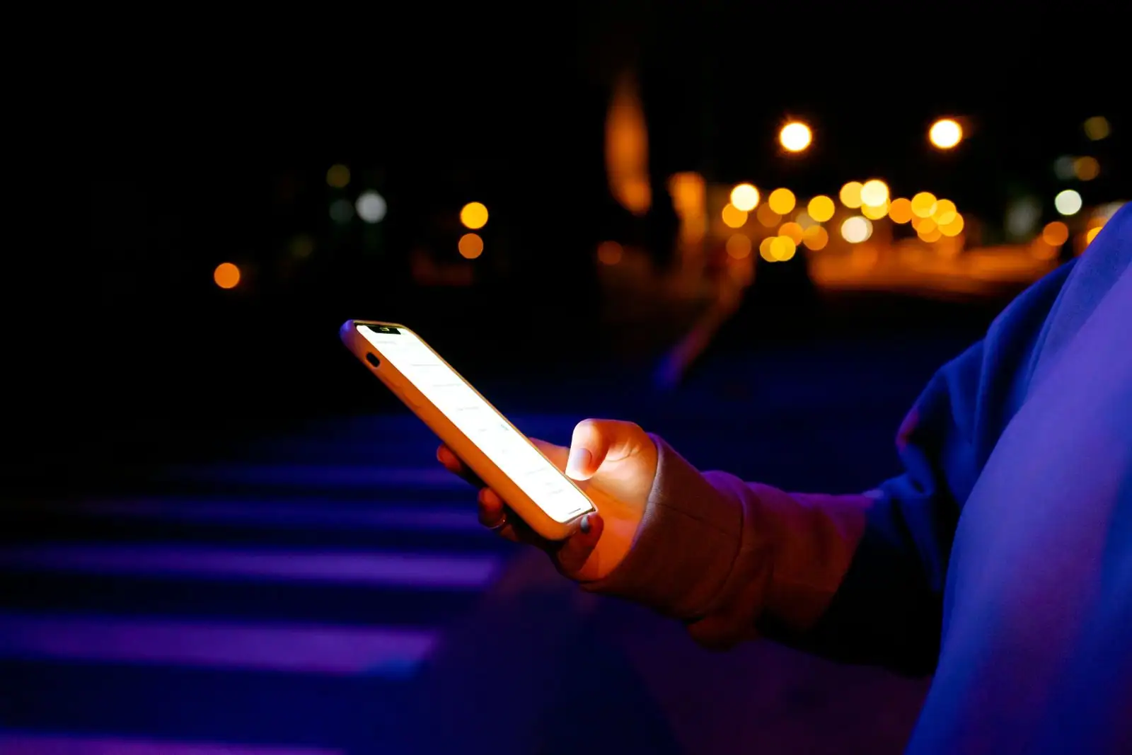 Main tenant un smartphone allumé la nuit en extérieur, lumières de la ville floues en arrière-plan.