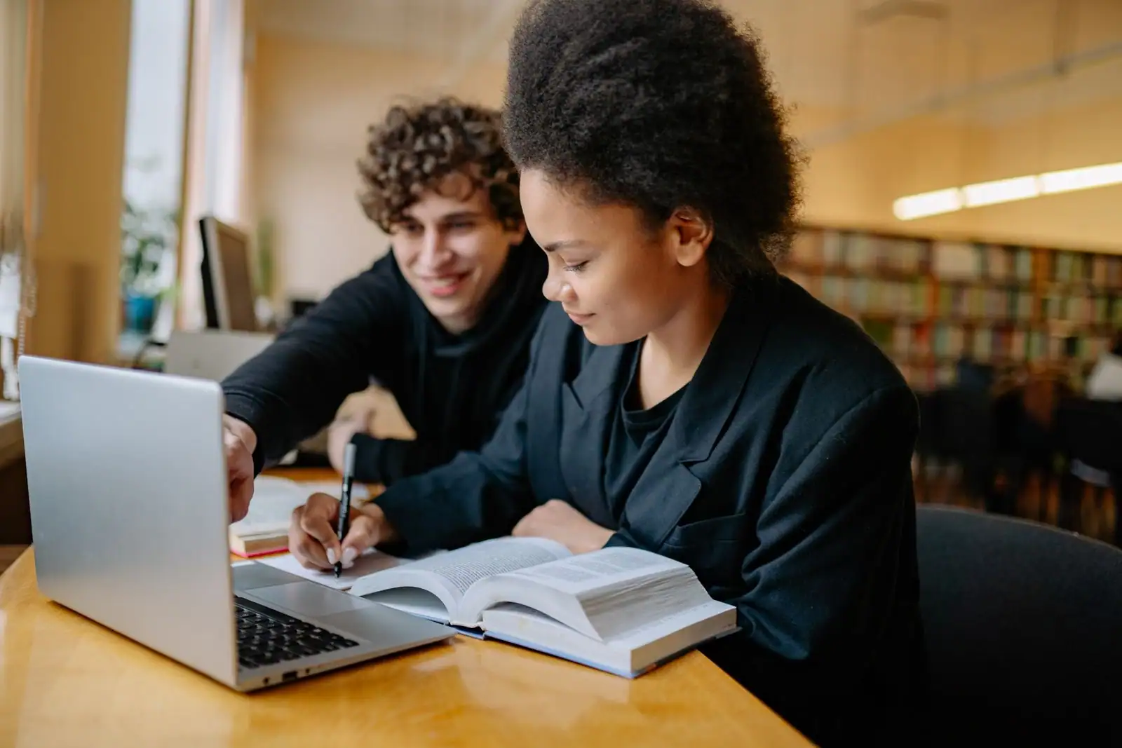 Deux étudiants attablés en bibliothèque avec un ordinateur portable, un livre ouvert et des notes