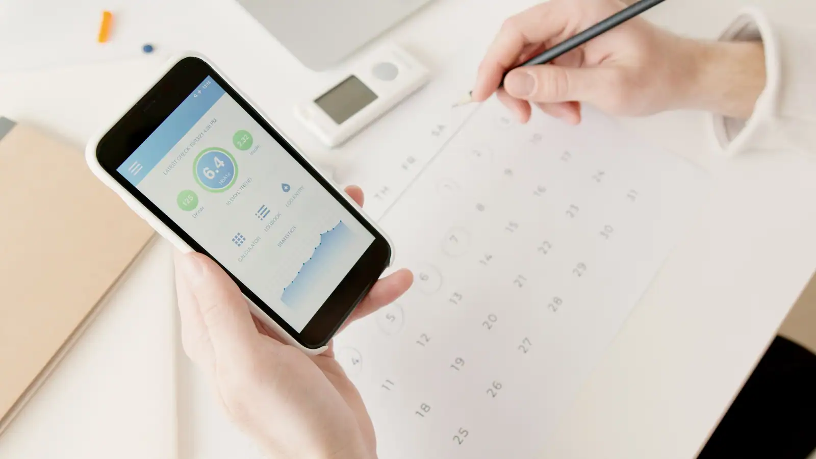 Main tenant un smartphone affichant une application de suivi santé, posé au-dessus d’un calendrier papier.