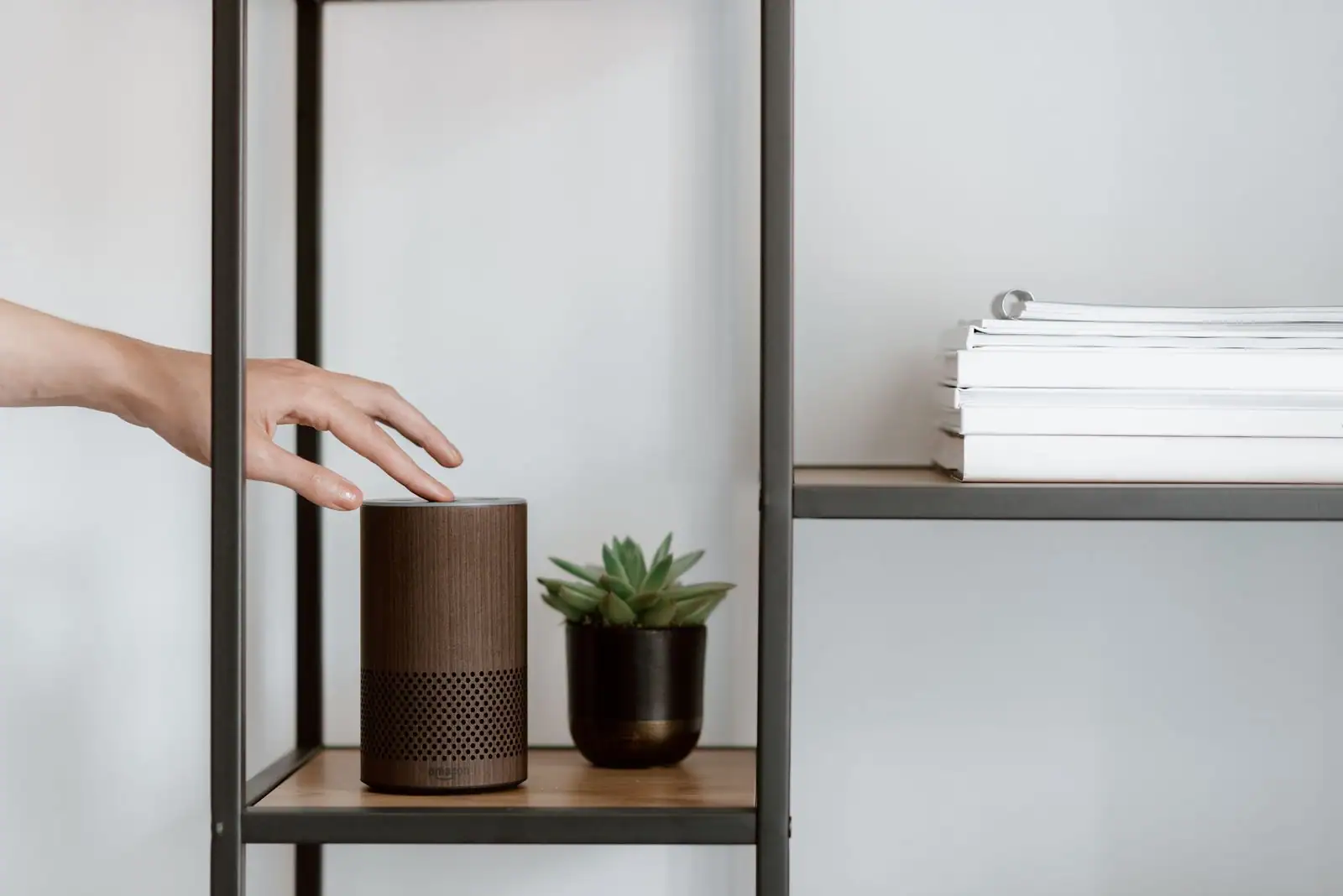 Main s'approchant d'une enceinte connectée à commande vocale posée sur une étagère.