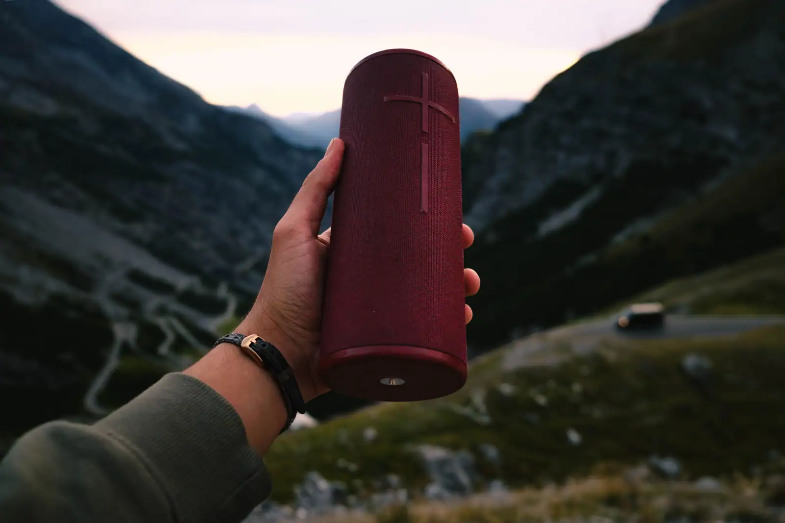 Enceinte portable cylindrique en tissu tenue à la main devant un paysage de montagne au crépuscule