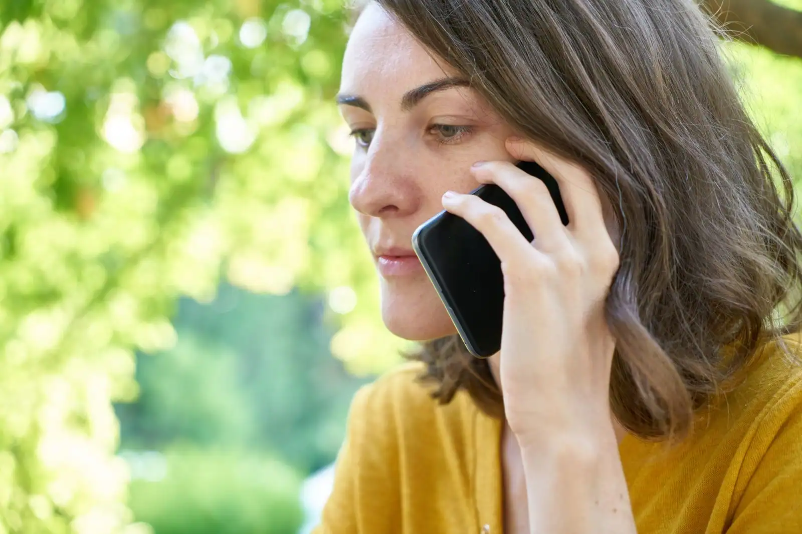 Femme passant un appel, smartphone tenu contre l'oreille, en extérieur devant un feuillage vert.