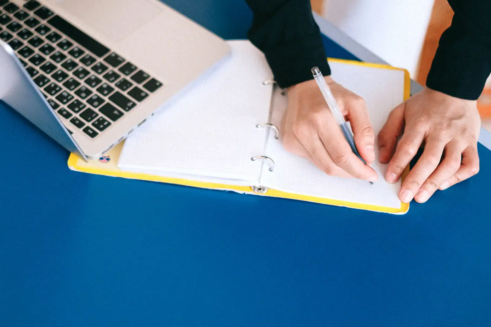 Mains en train d'écrire au stylo dans un carnet ouvert, à côté d'un ordinateur portable posé sur un bureau.