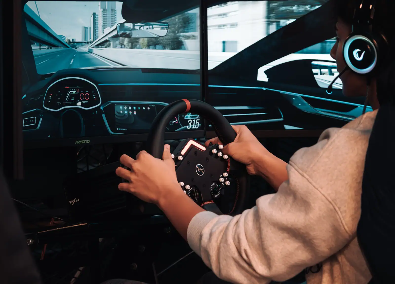 Joueur au casque tenant un volant de simulation devant trois écrans affichant une route urbaine.