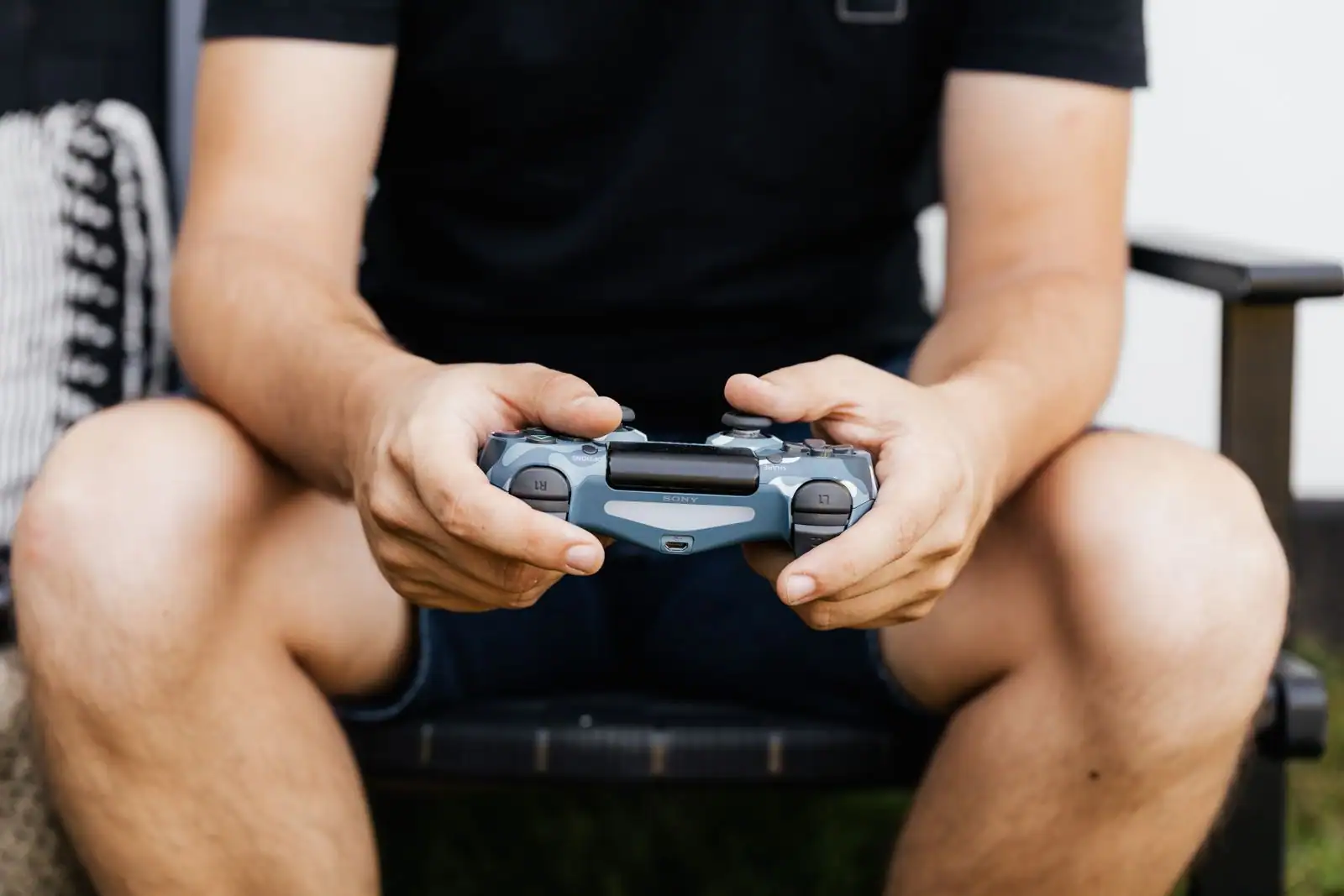 Personne assise en extérieur tenant à deux mains une manette de console PlayStation grise.