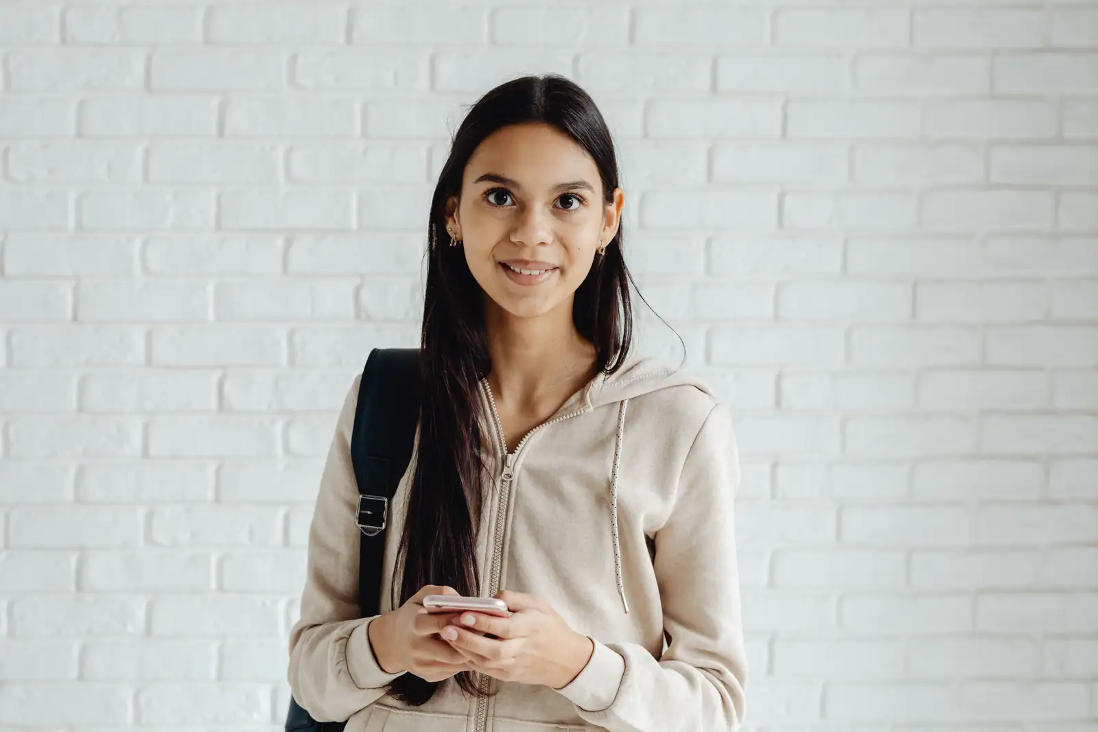 Jeune femme debout devant un mur de briques peint en blanc, son smartphone tenu à deux mains