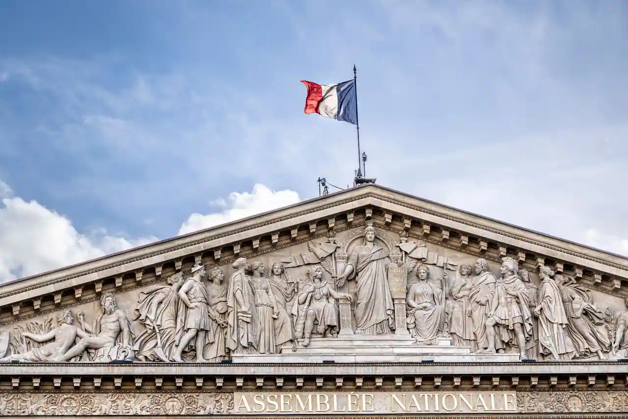 Fronton sculpté de l'Assemblée nationale à Paris, surmonté du drapeau français, sous un ciel nuageux.