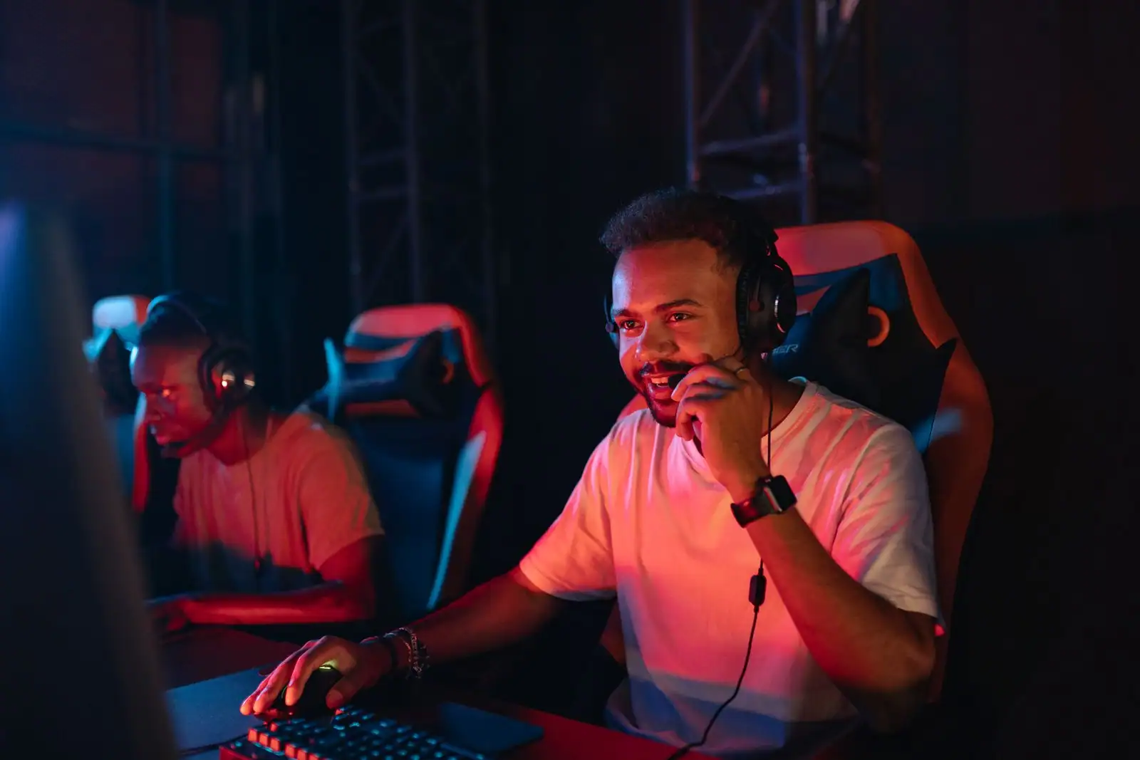 Deux joueurs équipés de casques-micros devant leurs écrans, dans une salle de jeu aux lumières rouges.