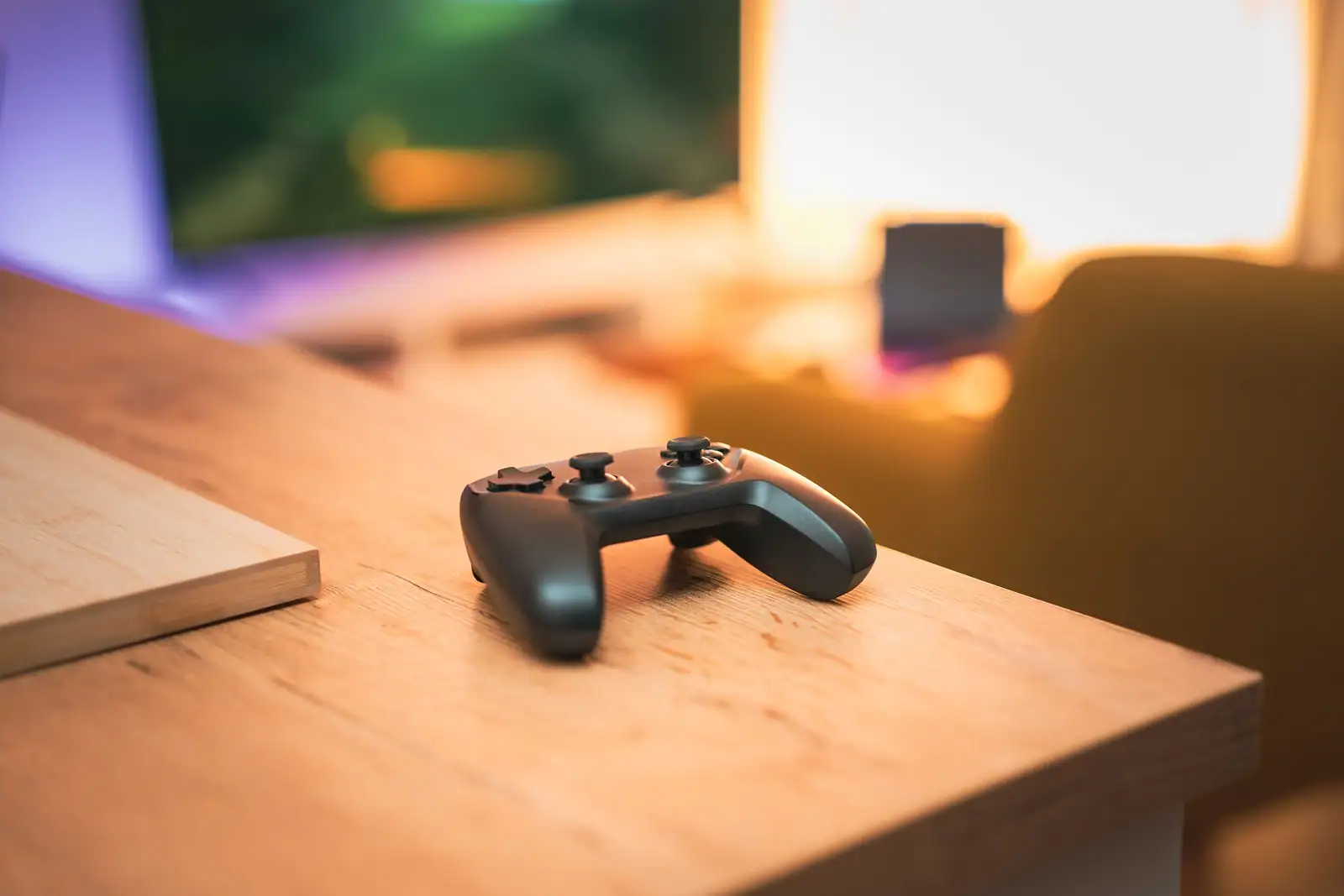Manette de jeu noire posée sur un bureau, devant un écran allumé dans une pièce éclairée en couleurs.