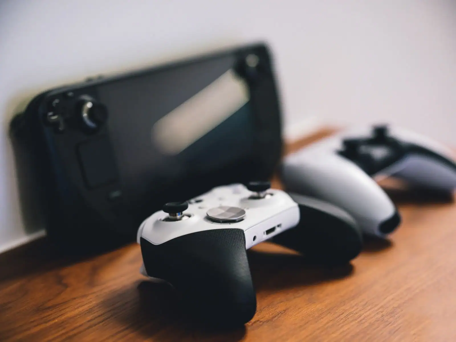 Manette de jeu blanche posée sur un bureau en bois, avec une console portable et d'autres manettes en arrière-plan.