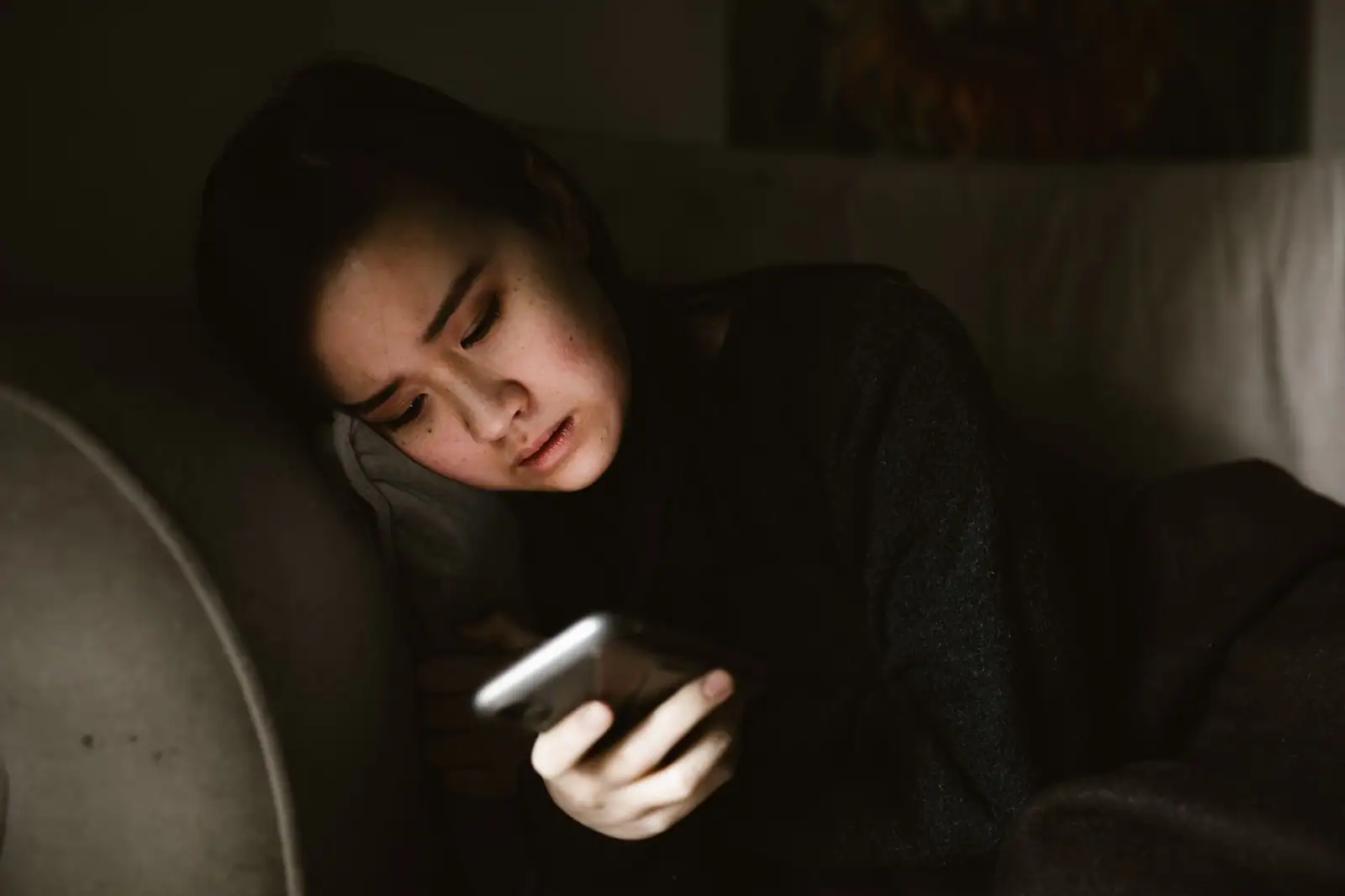 Jeune femme allongée qui regarde avec inquiétude l'écran lumineux de son téléphone.